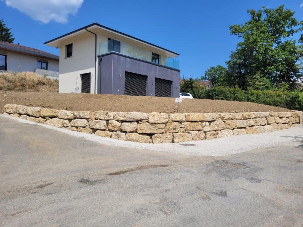 Maison moderne à deux étages sur une colline, avec mur de soutènement en pierre et route pavée sous un ciel bleu azur.