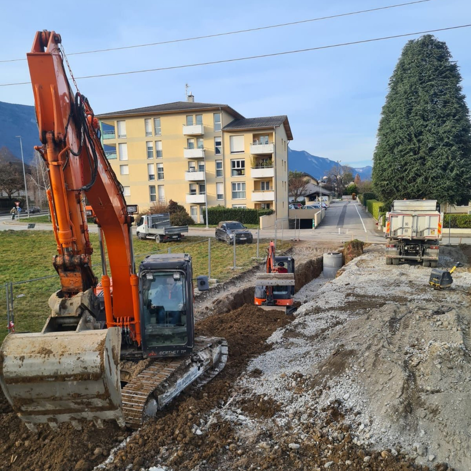 Une excavatrice creuse une tranchée boueuse sur un chantier routier à proximité d'immeubles d'habitation.