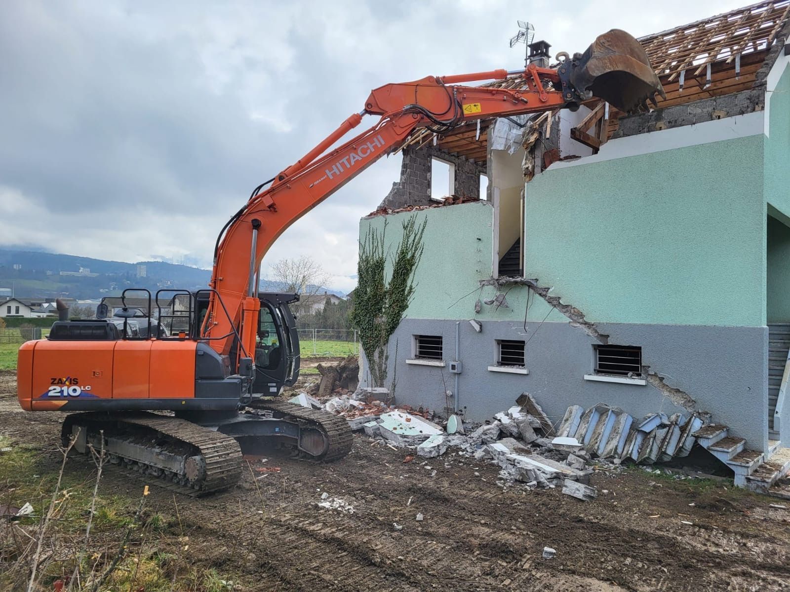 Une pelleteuse orange démolit un bâtiment endommagé à côté d'un mur vert menthe, des décombres jonchent le sol.