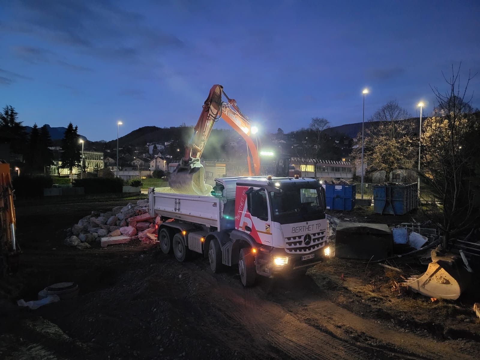 Une excavatrice charge un camion plateau au crépuscule sur un chantier de construction, avec des lumières et des montagnes en arrière-plan.