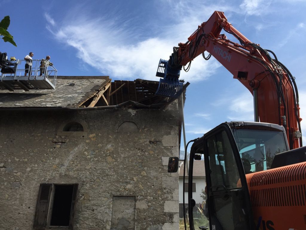 Une pelleteuse orange démolit le toit d'un bâtiment en pierre sous un ciel bleu.