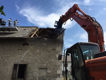 Une pelleteuse rouge démolit le toit d'un bâtiment en pierre sous un ciel bleu ; on aperçoit des ouvriers sur le dessus.