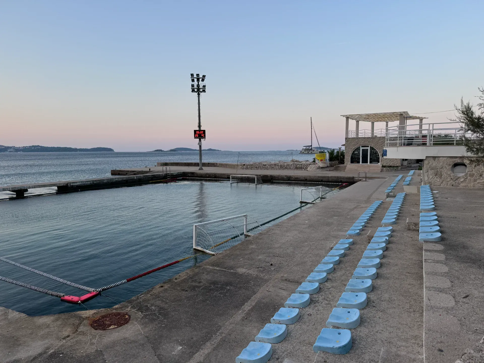 An outdoor water polo pool by the sea with rows of blue spectator seats at dusk.