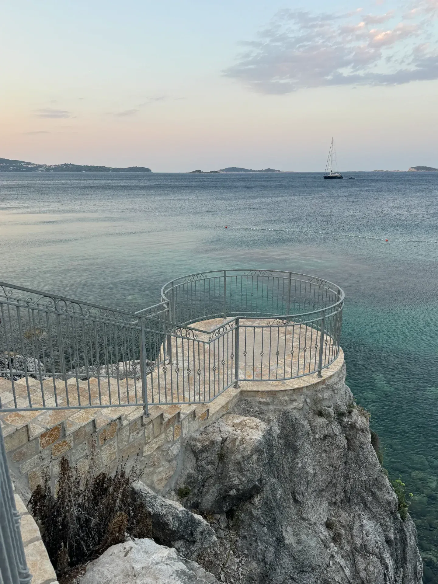 A stone viewing platform with a metal railing overlooks the calm, blue sea under a soft, dusk sky with a distant sailboat.
