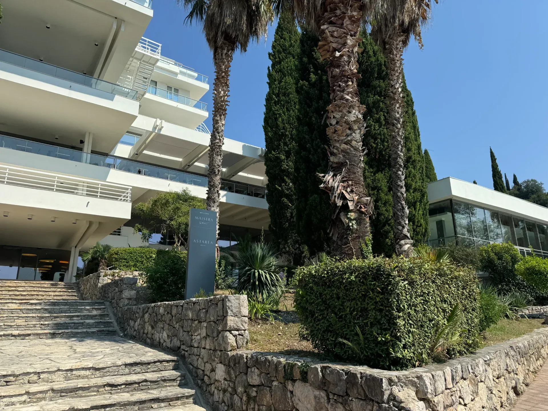 Modern white hotel with tiered balconies and stone steps, surrounded by palm trees and lush landscaping under a clear sky.