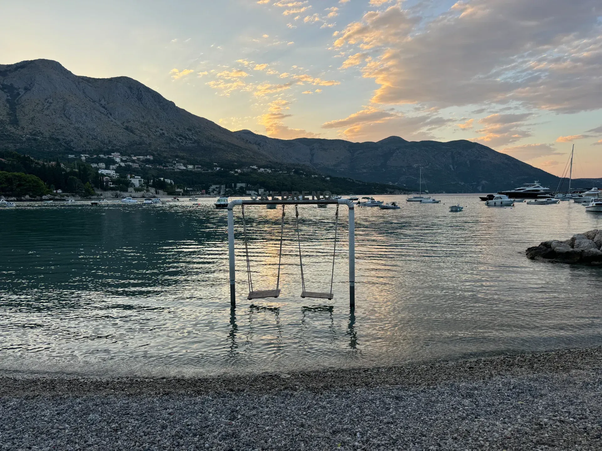 A set of swings standing in the shallow water of a calm bay at sunset, with mountains and boats in the distance.