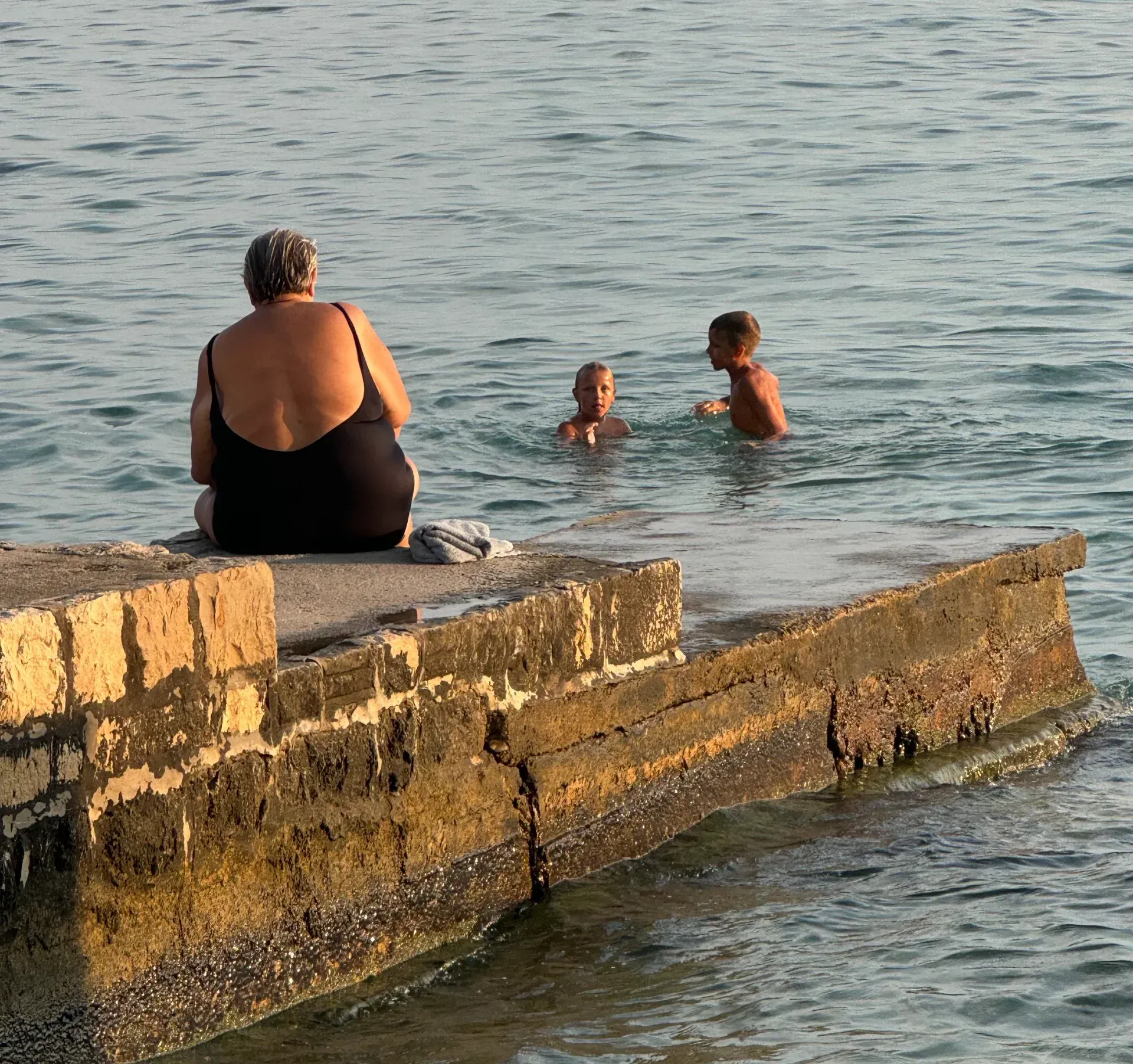 A person in a black swimsuit sits on a concrete pier watching two people swim in the ocean.