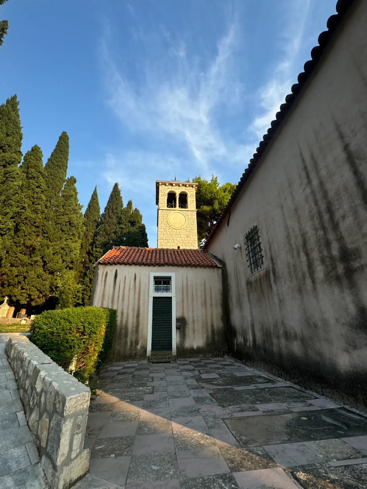 Stone church and bell tower with tile roof and cypress trees against a blue sky, viewed from a stone courtyard.