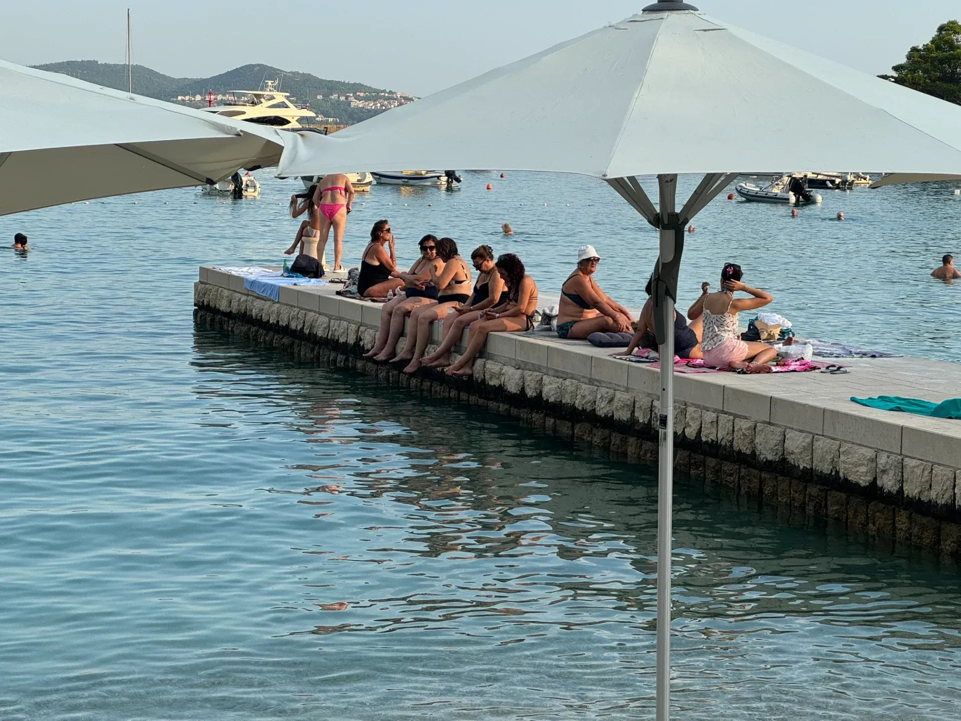 People sit on a concrete pier by the water under a large sun umbrella on a sunny day with boats in the distance.