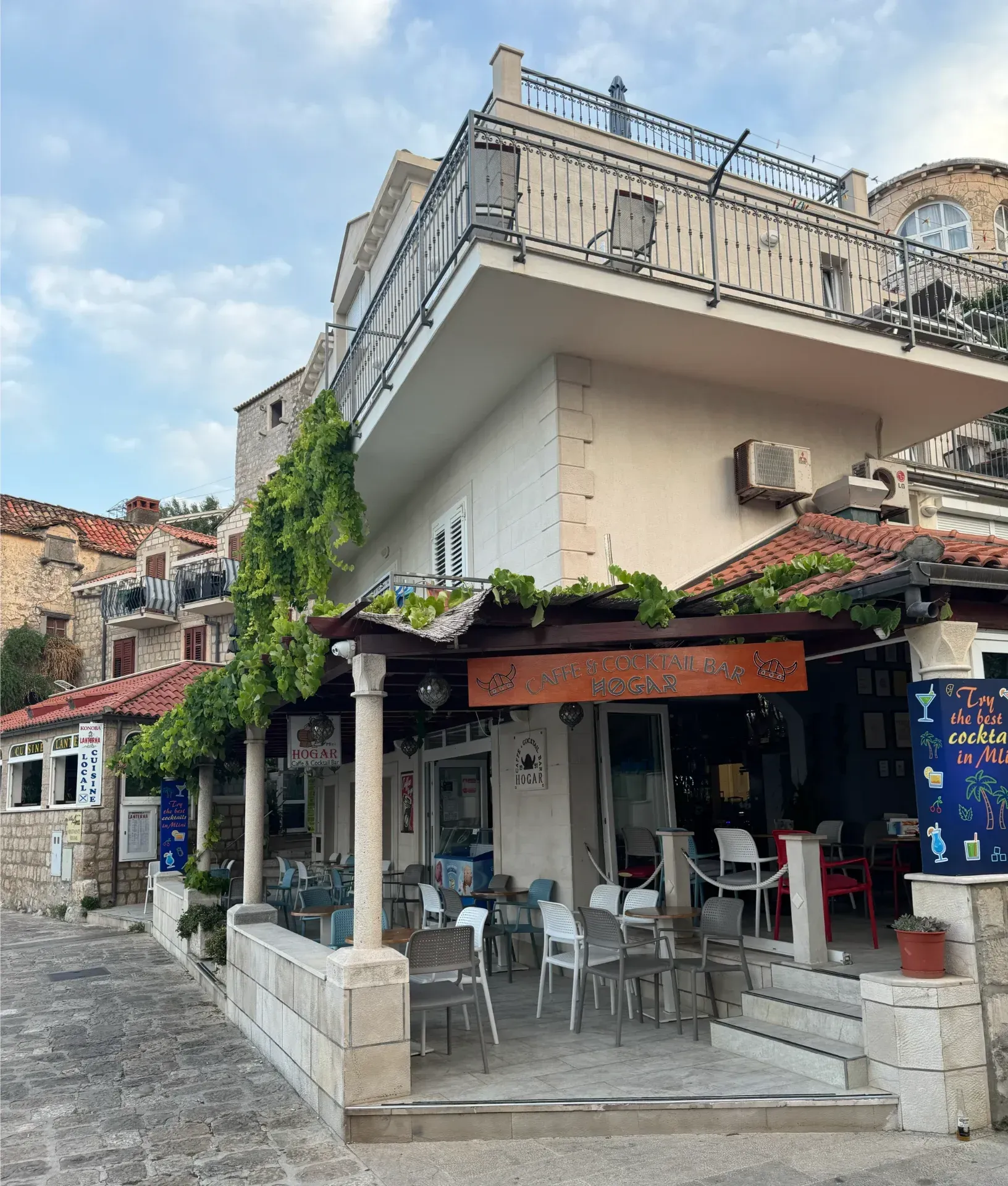 A two-story building with a stone-paved patio, outdoor cafe seating, and green vines growing over a wooden trellis.