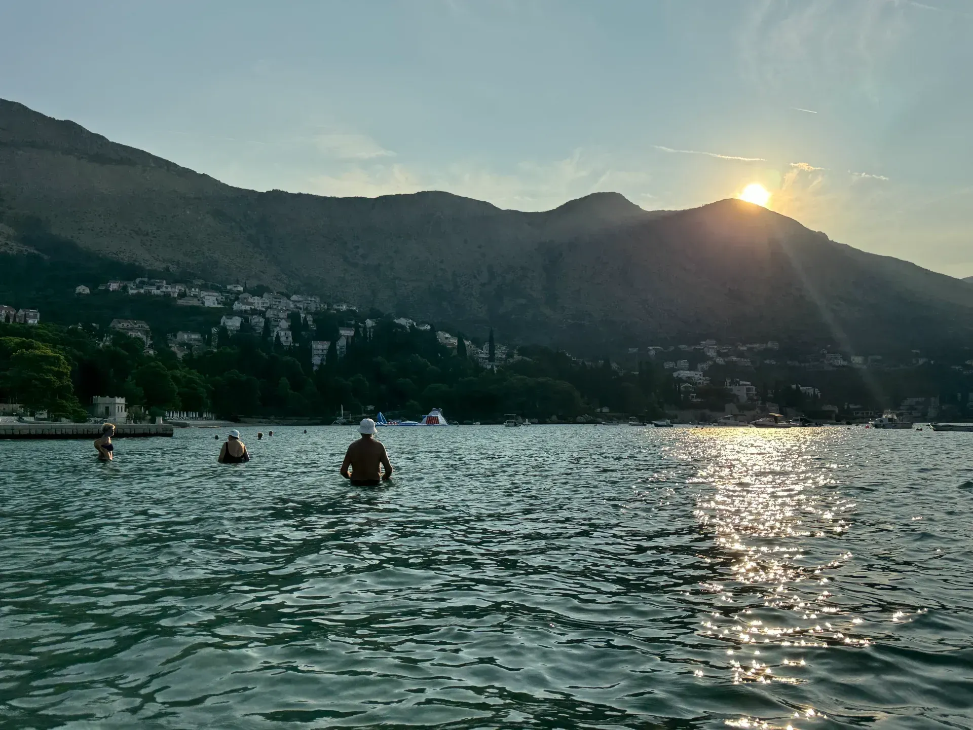 People wade in a calm, sunlit sea before mountains at sunset, with light shimmering on the water's surface.