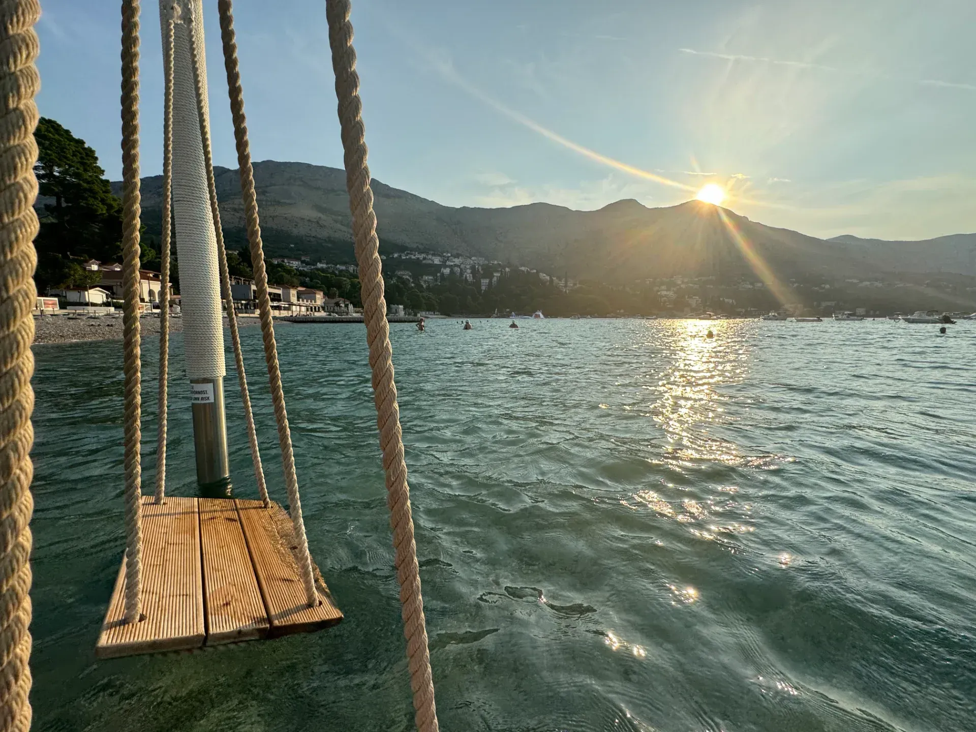 A wooden swing hangs over calm water at sunset, with distant mountains and a shoreline visible in the background.