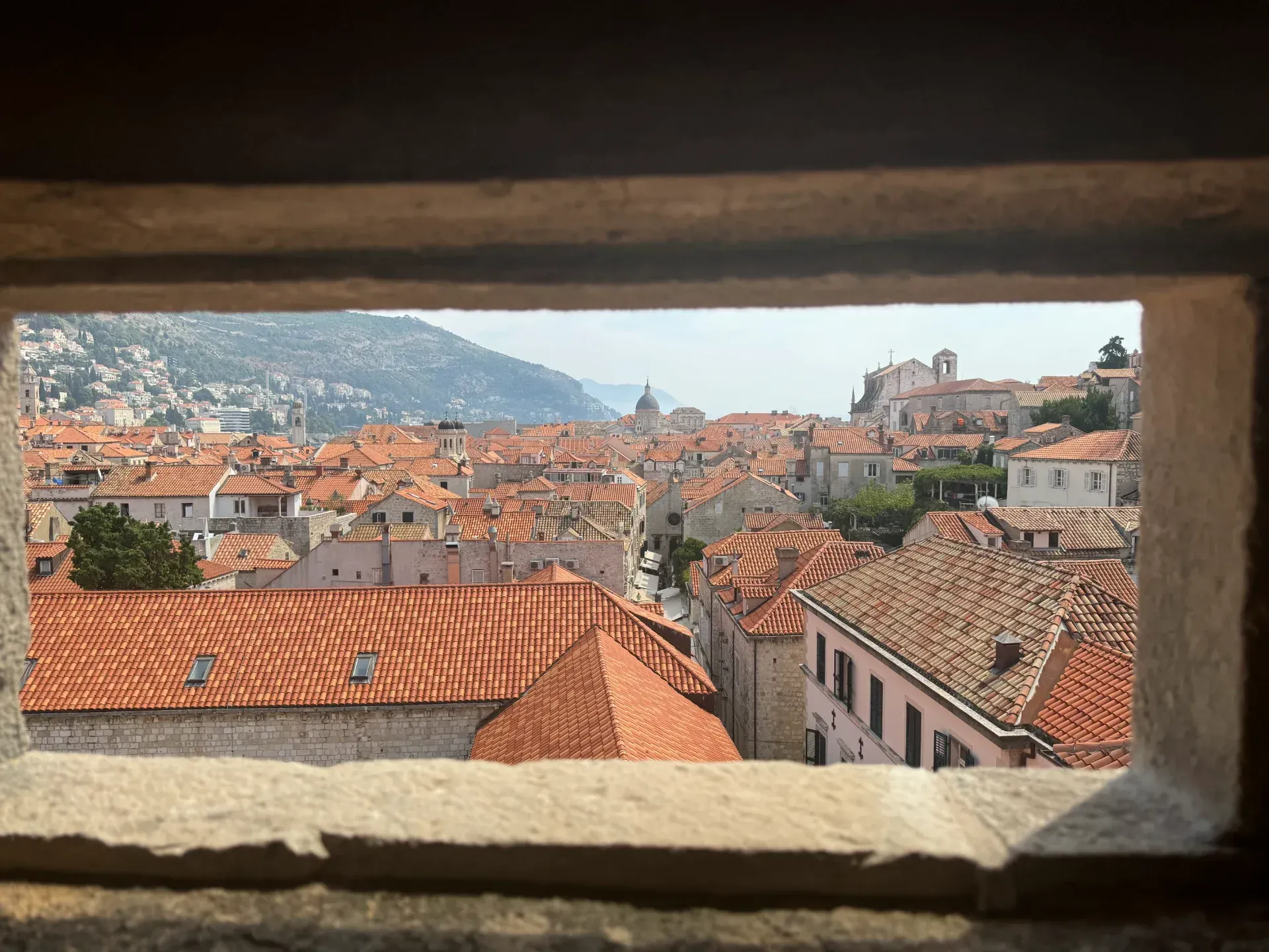 A stone window frame overlooks the terracotta-tiled rooftops of Dubrovnik’s historic old town near the sea.