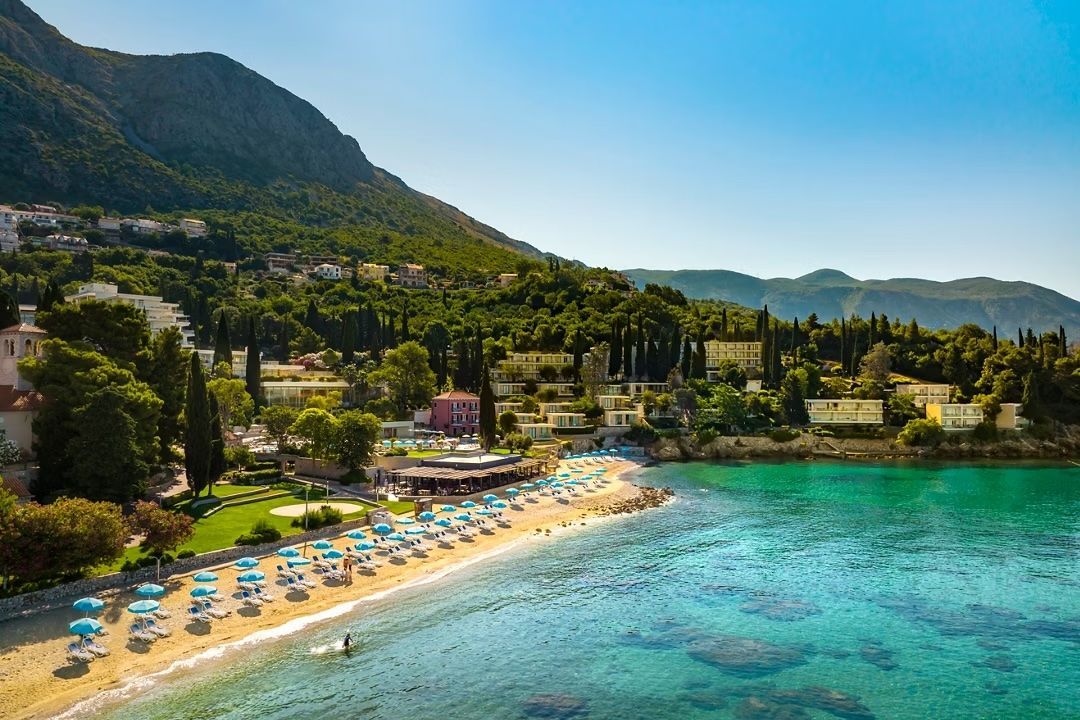 An aerial view of a turquoise beach with rows of blue umbrellas, nestled against a tree-covered hillside.