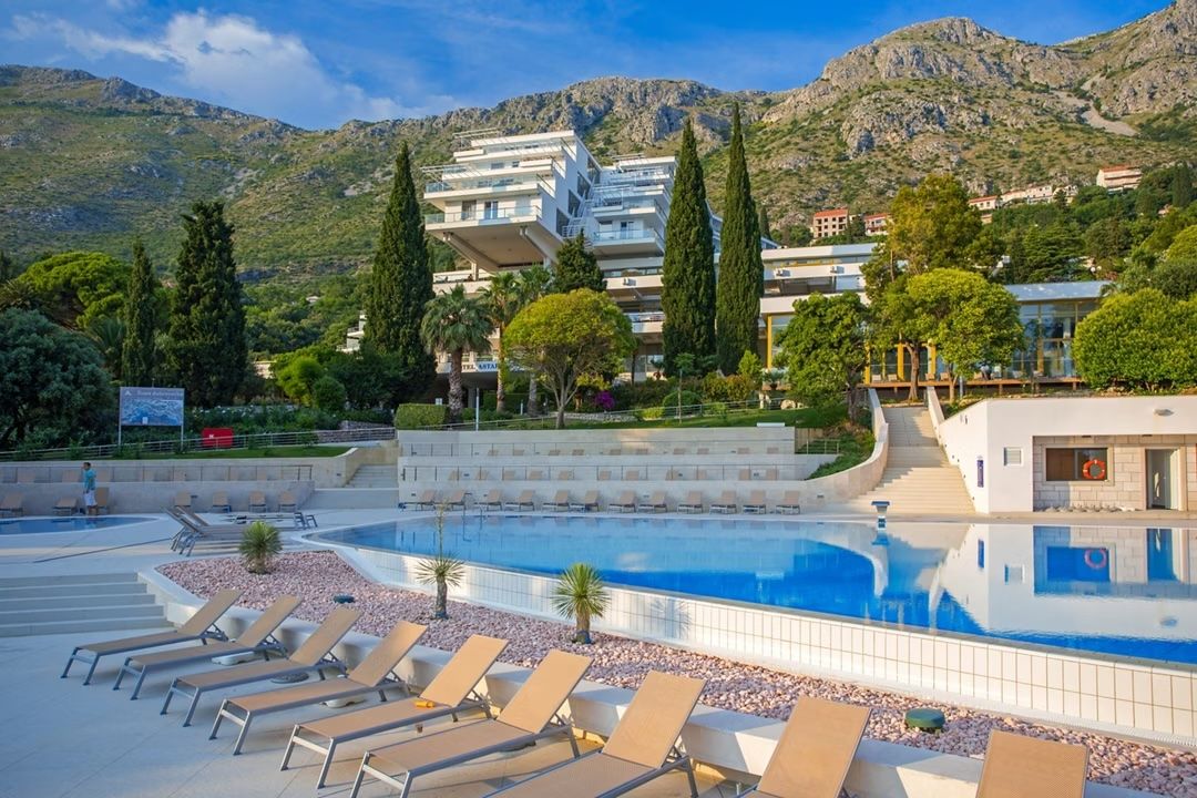 A multi-story Hotel Astarea in Croatia with a tiered exterior stands before mountains, overlooking a swimming pool with lounge chairs.