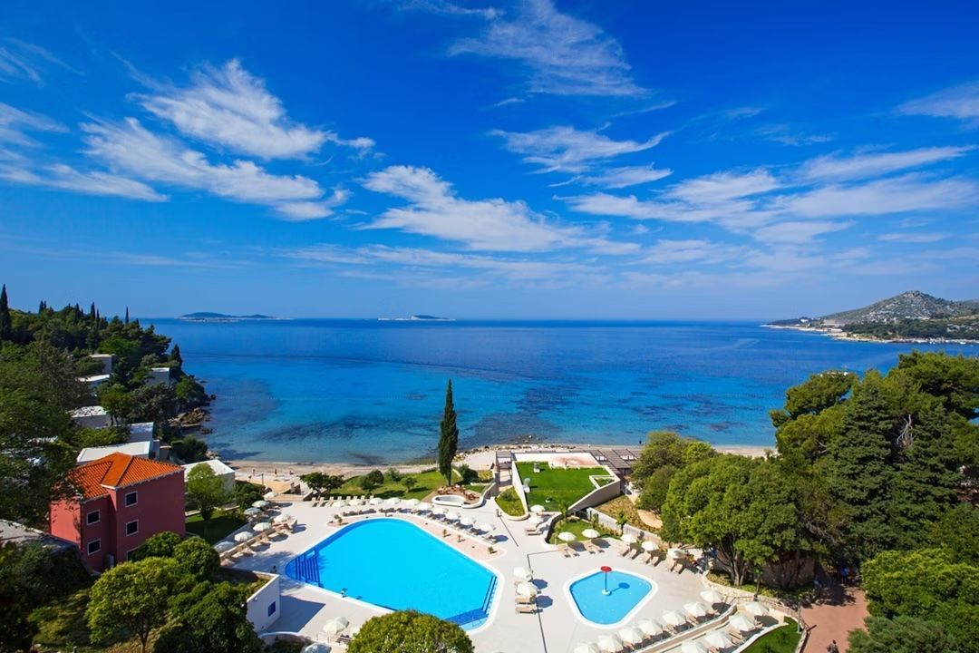 An aerial view of a resort pool area next to a sandy beach and blue sea under a bright, sunny sky.