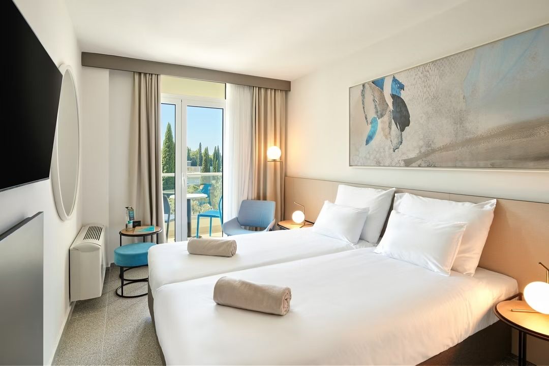 A modern hotel room with two white twin beds, beige curtains, a balcony, wall art, and warm lighting.