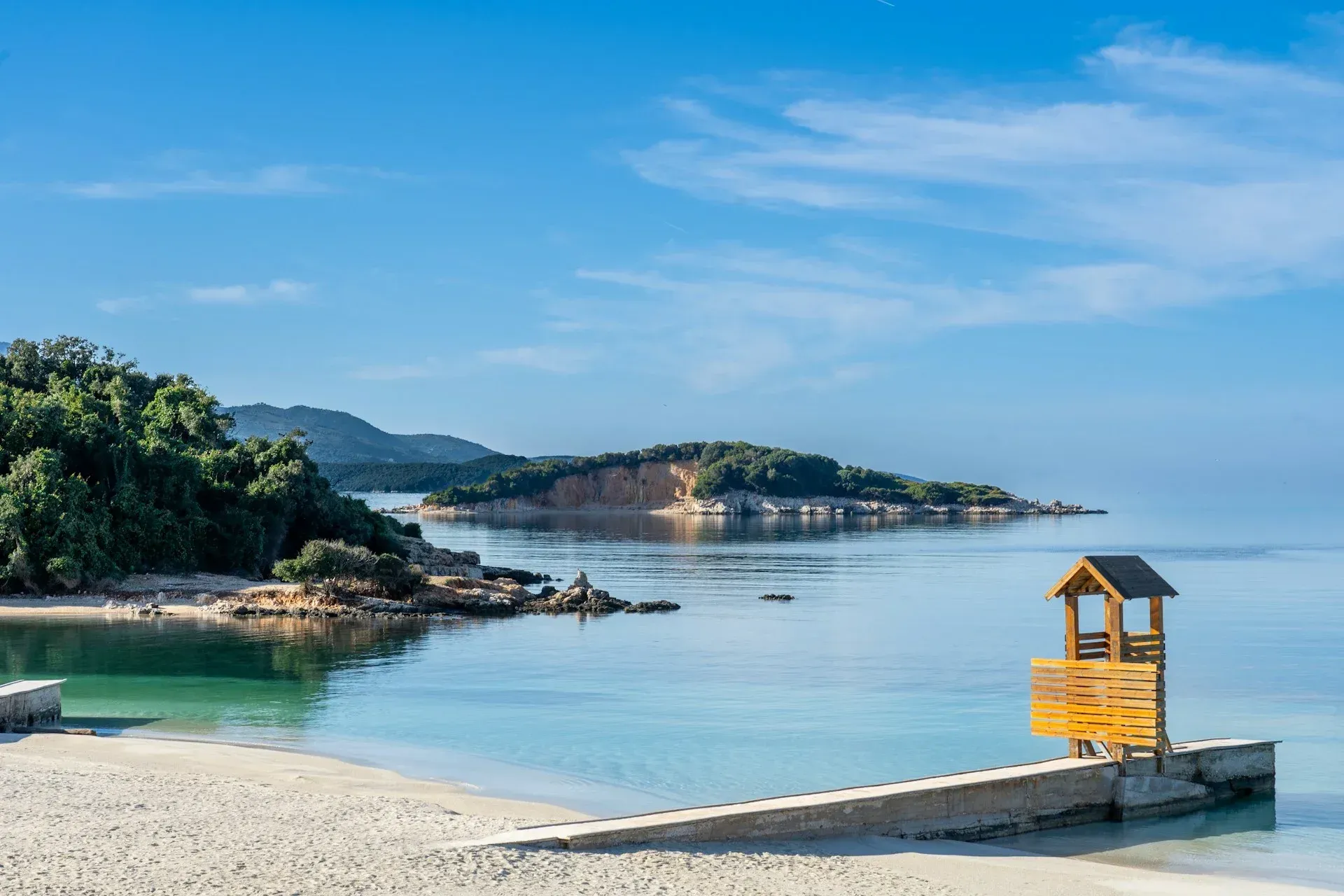 A wooden lifeguard tower stands on a pier overlooking a calm, turquoise beach with a lush, hilly island in the distance.