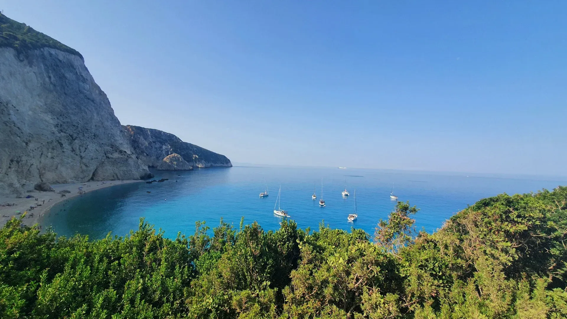 A scenic view of a turquoise bay with sailboats, bordered by a large rocky cliff on the left and lush green trees below.