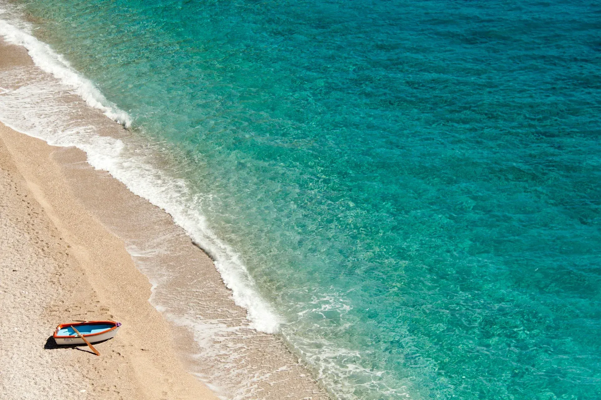 A lone, small boat rests on a pebble beach beside clear, turquoise ocean water with gentle waves lapping the shore.