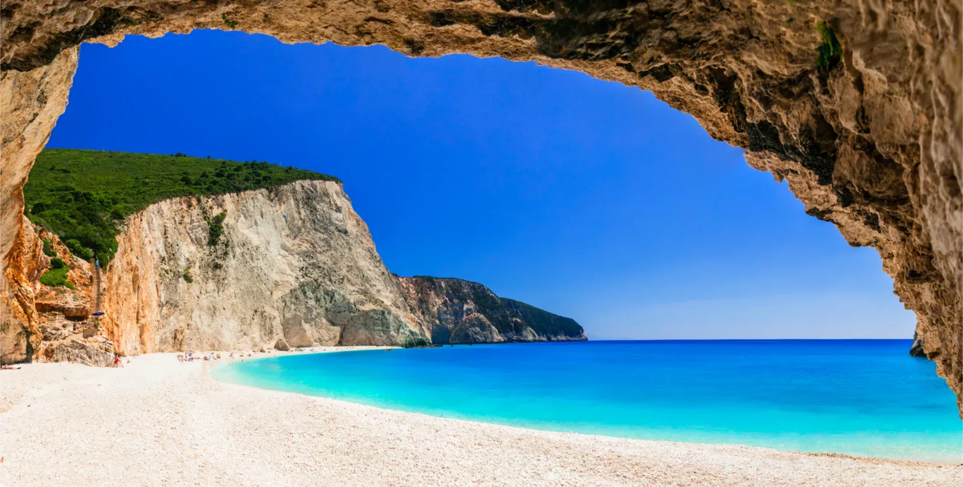 Steep white cliffs rise above a crescent-shaped beach with turquoise water and small boats along the coast of Lefkada.