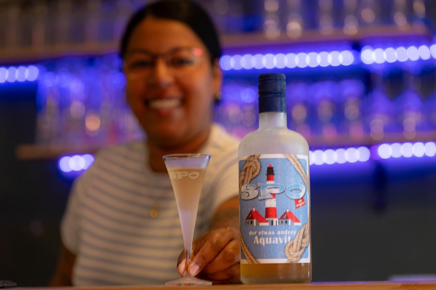 Eine Frau steht hinter einer Bar und hält ein Martiniglas und eine Flasche Alkohol in der Hand.