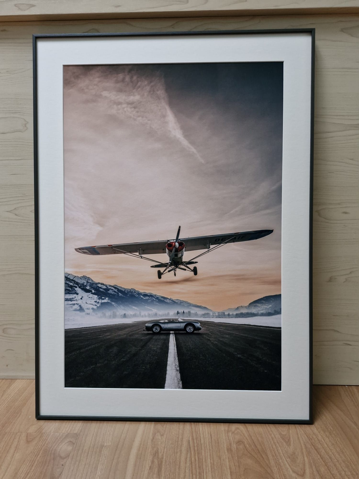 Gerahmter Druck eines Sportwagens auf einer Straße unter einem tief fliegenden Flugzeug, im Hintergrund Berge.