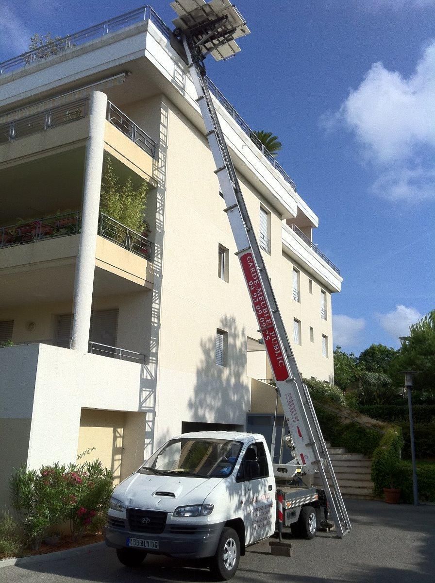 Un camion blanc équipé d'une échelle transporte des objets vers un bâtiment à la façade blanche, sous un ciel bleu.