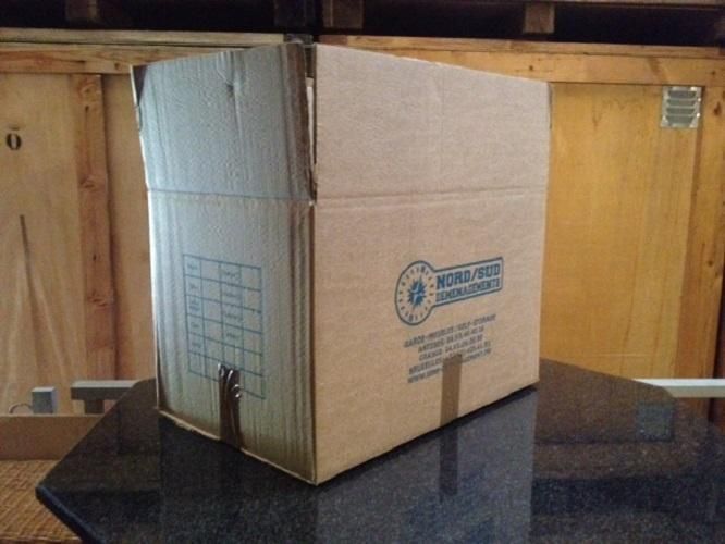 Boîte en carton posée sur une surface réfléchissante, portant une étiquette et un emblème bleu.