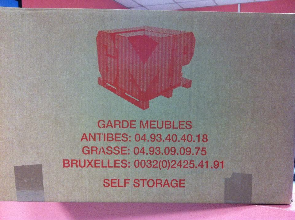 Boîte en carton avec texte rouge et illustration. Indique les lieux de stockage : Antibes, Grasse et Bruxelles.