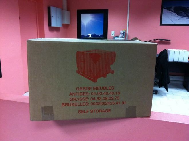 Grande boîte en carton avec logo et texte rouges, à l'intérieur d'une pièce rose.