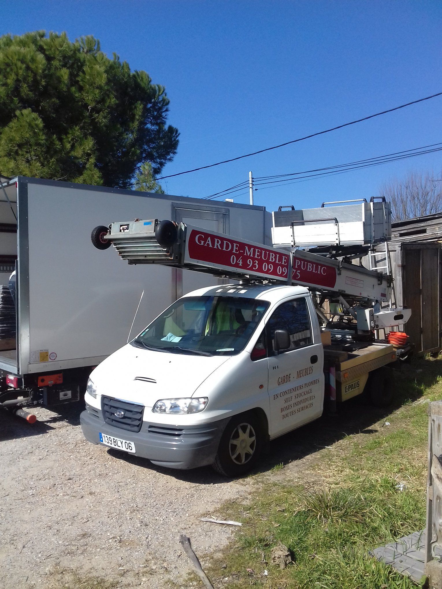 Camion blanc avec nacelle élévatrice, stationné près d'un camion cargo blanc. Journée ensoleillée, ciel bleu.