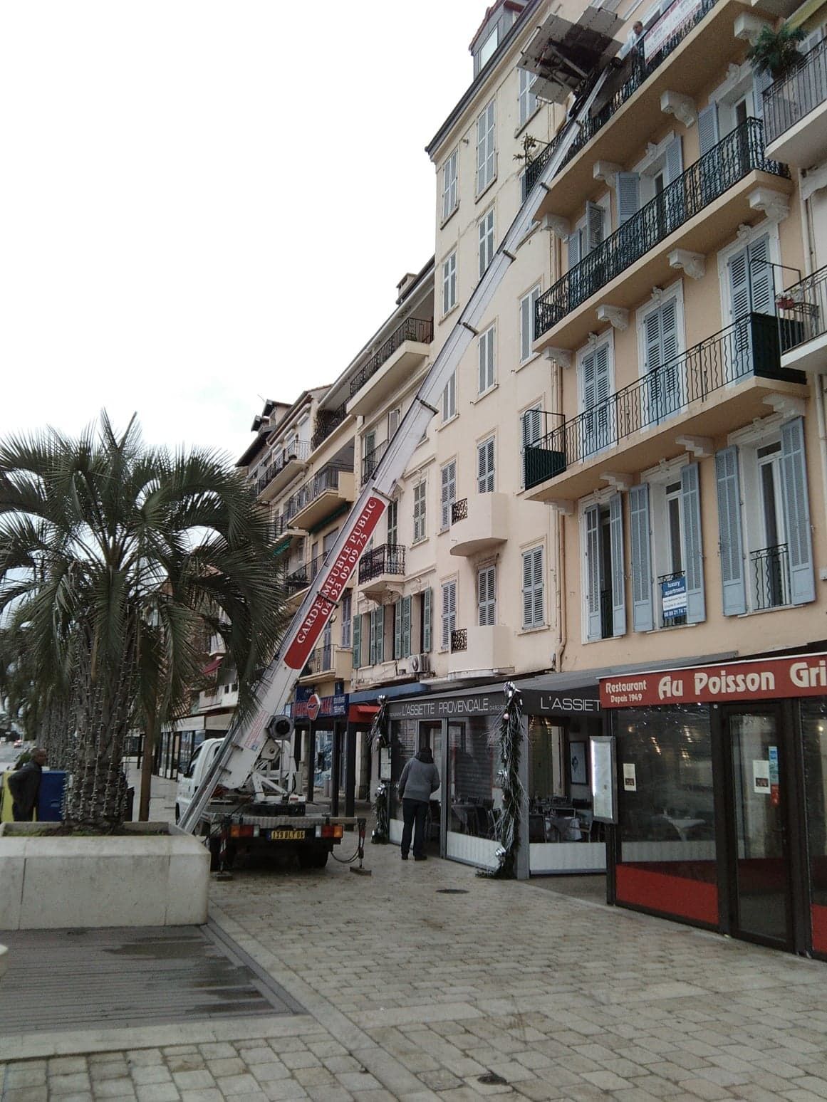 Un camion à hayon élévateur est amarré contre un bâtiment beige aux volets ouverts, probablement pour un déménagement.