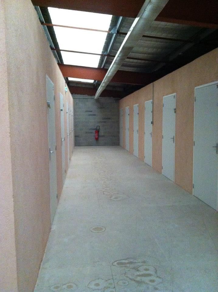 Couloir d'accès à l'unité de stockage, sol en béton, murs beiges, portes blanches, puits de lumière, extincteur au fond.