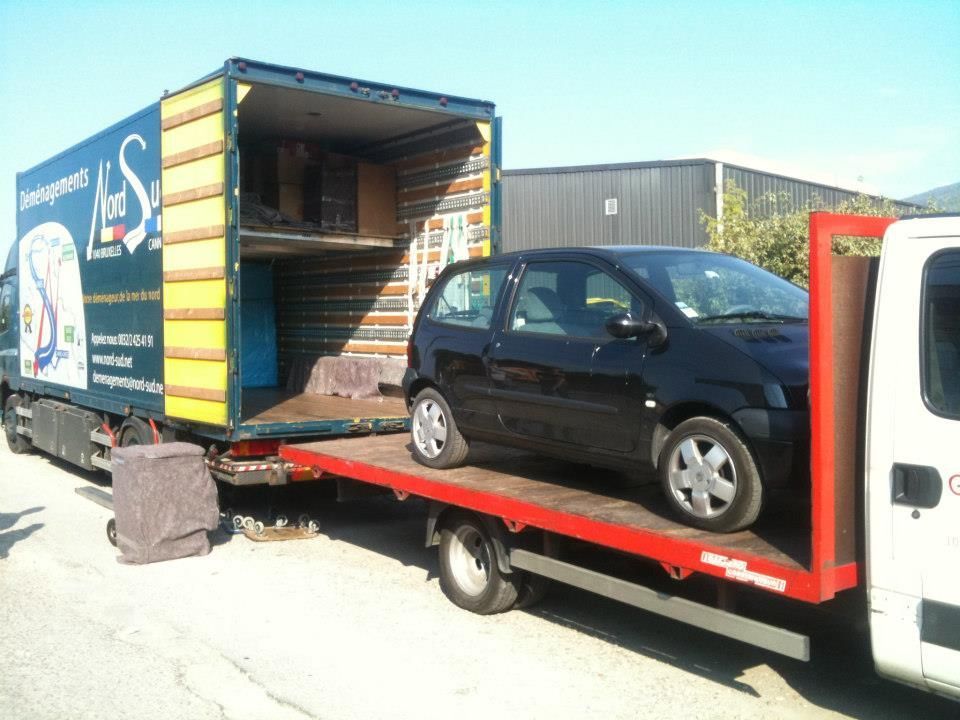 Voiture noire sur une dépanneuse à plateau, à côté d'un camion de déménagement, en extérieur par une journée ensoleillée.