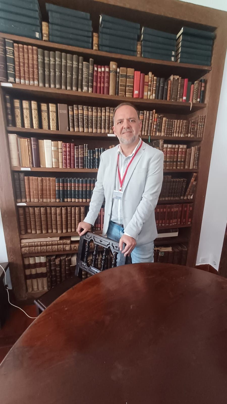 Un hombre con una chaqueta ligera está de pie frente a una estantería. Sonríe, con las manos apoyadas en una silla, en una biblioteca.