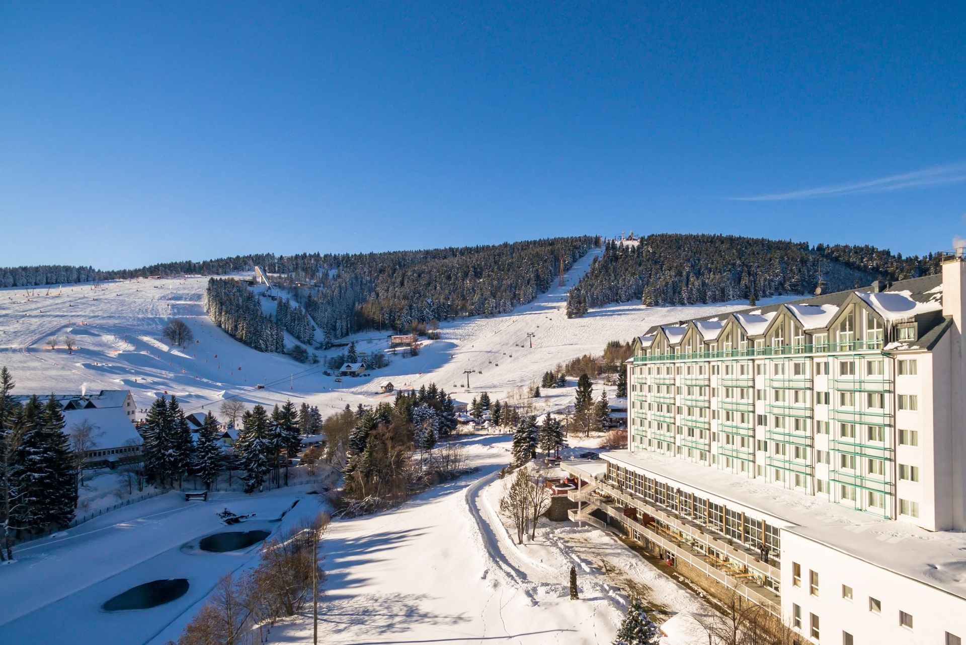 Verschneites Skigebiet mit Pisten, Kiefern und einem großen Hotel unter klarem, blauem Himmel