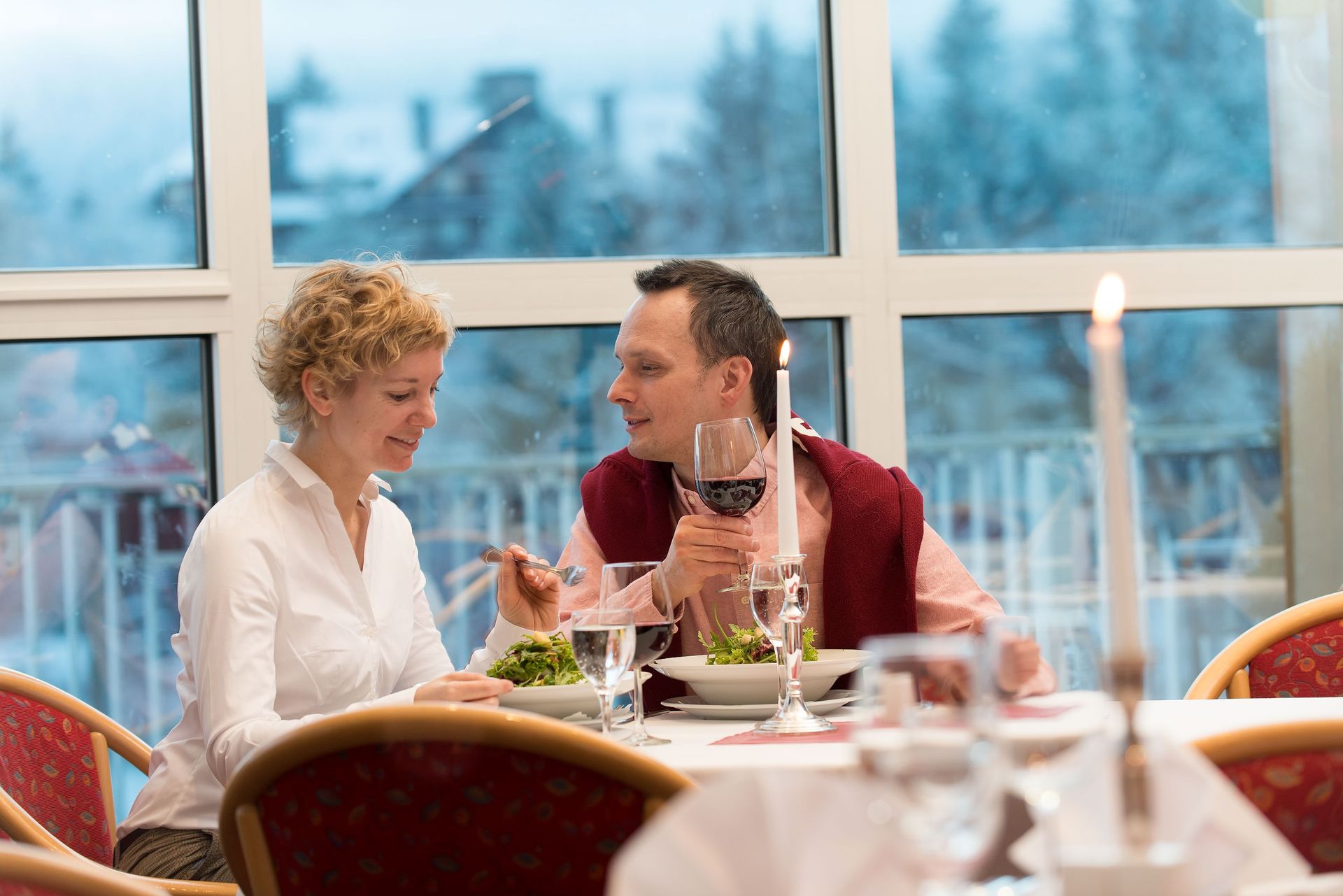 Paar an einem Restauranttisch, Mann hält Weinglas, lächelt, Frau schaut zu, helles Fenster.