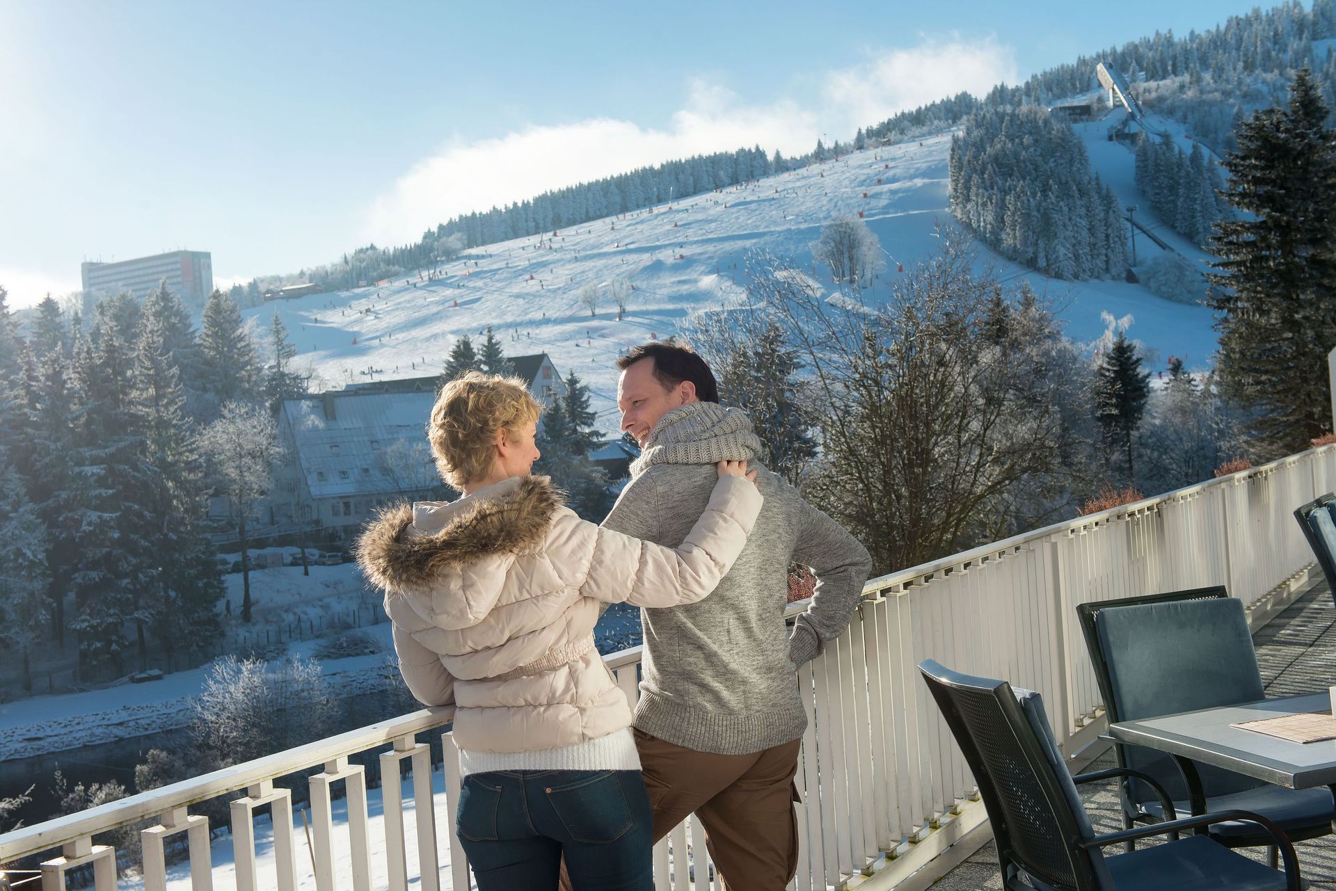 Ein Paar in Winterjacken auf der verschneiten Terrasse, Blick auf sonnenbeschienene Skipisten und Berge