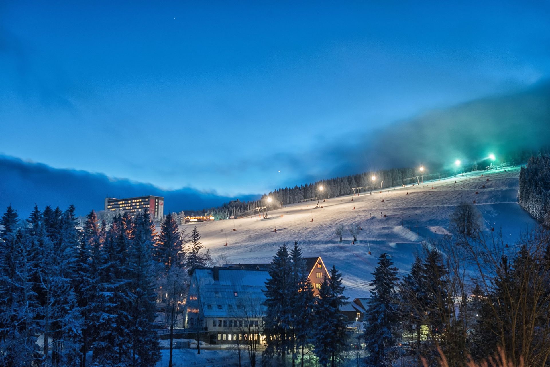Verschneite Skipiste in der Abenddämmerung mit beleuchteten Pisten, Kiefern und Chalets unter blauem Abendhimmel