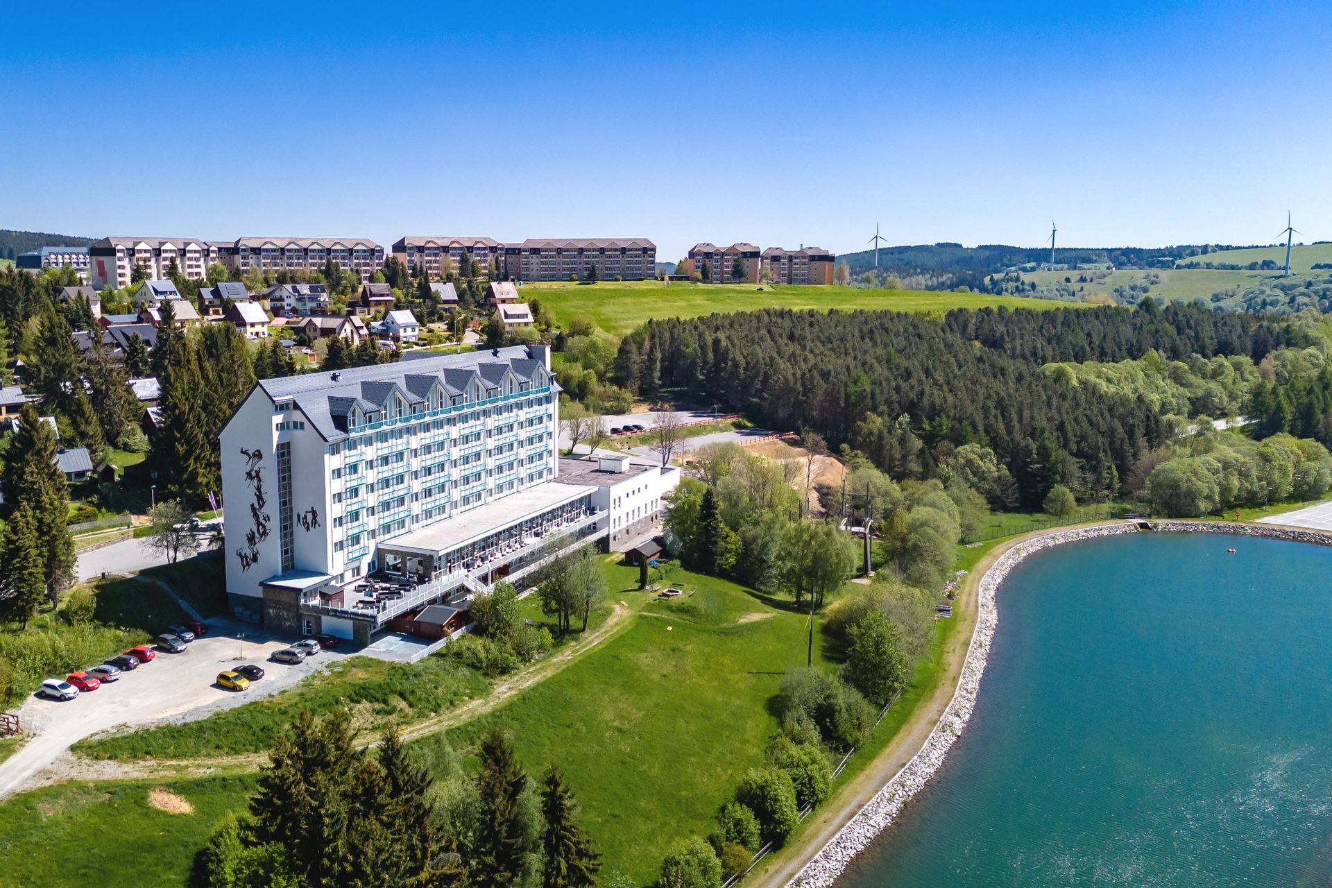 Luftaufnahme eines modernen weißen Hotels an einem blauen See, umgeben von grünen Rasenflächen und Bäumen