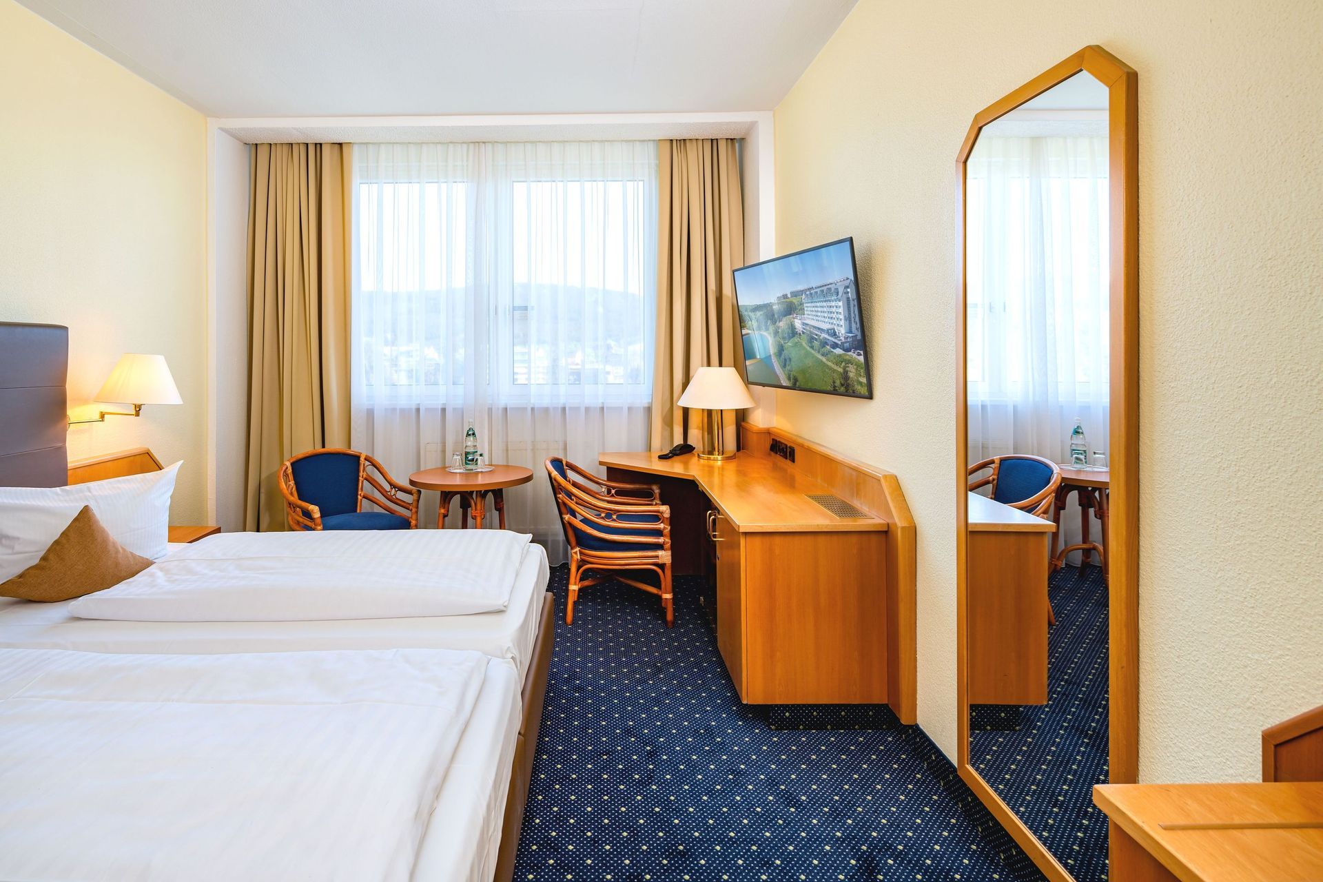 Hotelzimmer mit zwei Einzelbetten, Schreibtisch, Stuhl, Wandfernseher und großem Fenster mit Vorhängen
