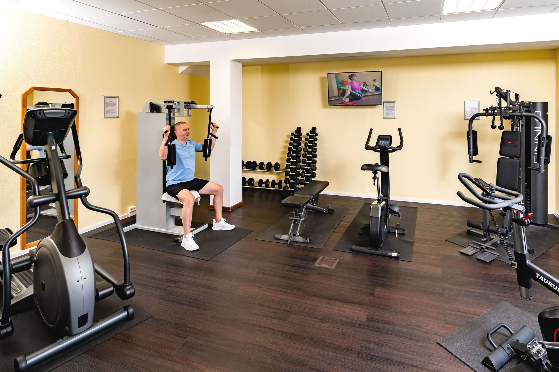 Innenraum eines Fitnessstudios mit Trainingsgeräten und einer Person, die an einer sitzenden Krafttrainingsmaschine trainiert.