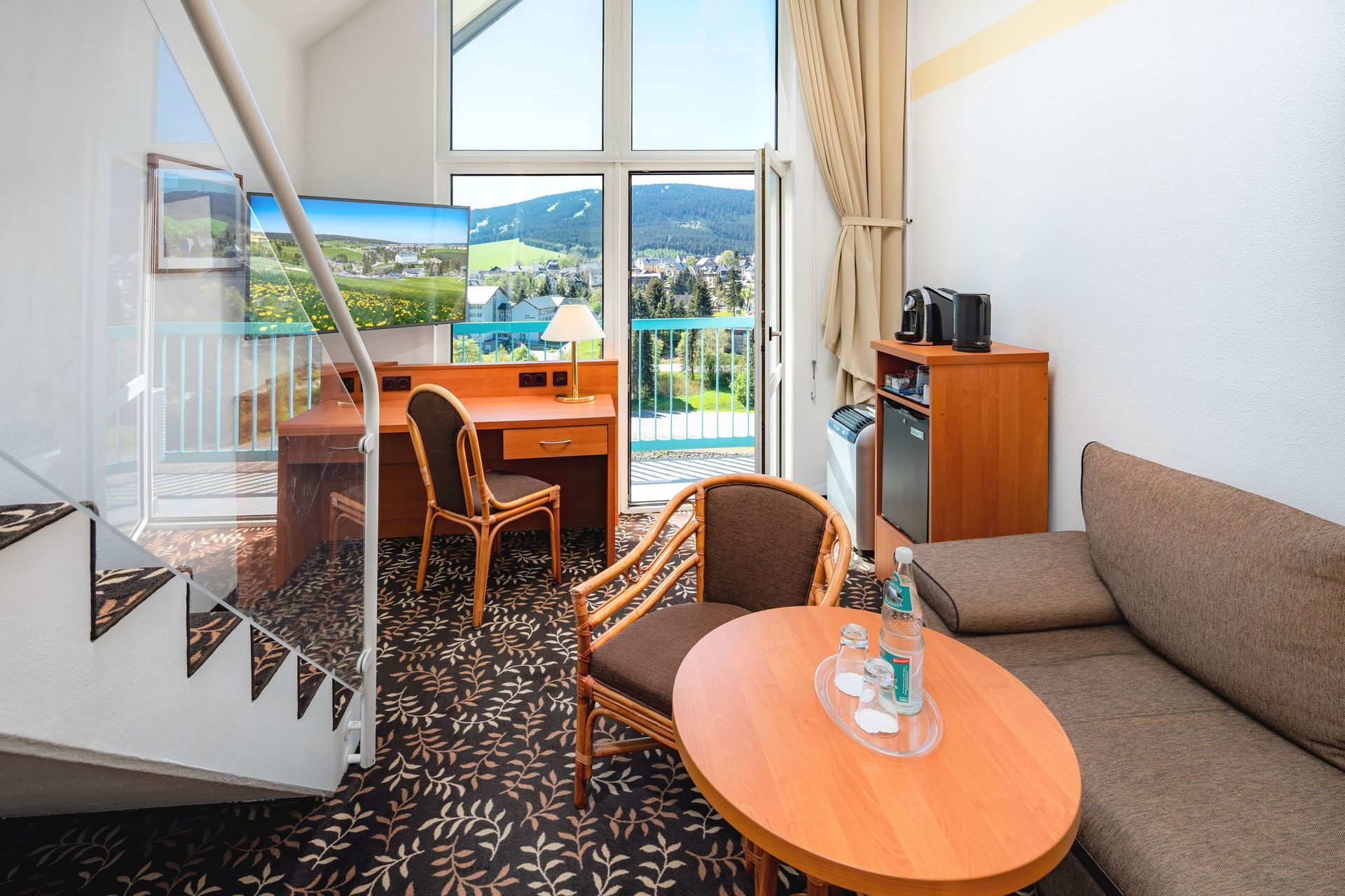 Gemütliches Hotelzimmer mit Schreibtisch, Sofa, rundem Tisch und großen Fenstern mit Blick auf einen See und die Berge