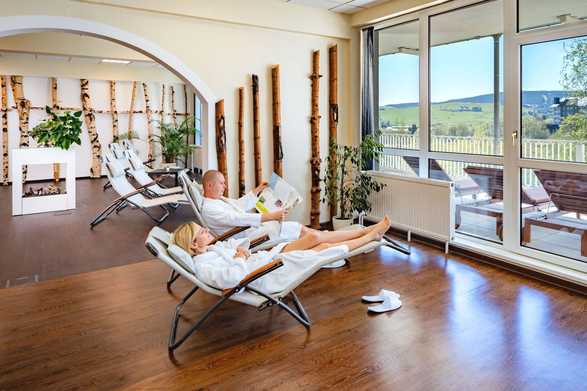 Zwei Personen entspannen auf Liegestühlen in einem hellen Wellnessraum mit großen Fenstern und malerischer Aussicht.