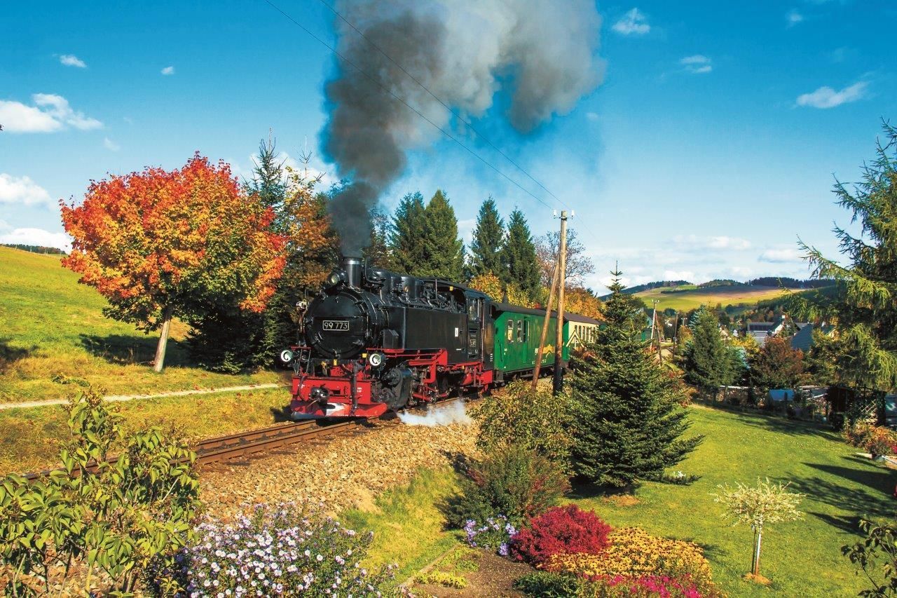 Eine Dampflokomotive zieht grüne Waggons durch eine farbenprächtige Herbstlandschaft, über der Rauchwolken aufsteigen.