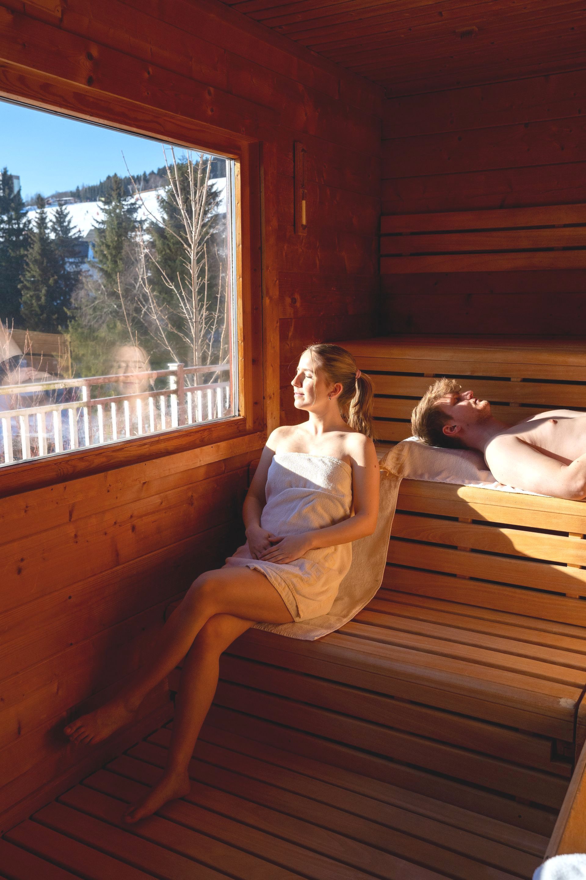 Eine Frau sitzt in einer Sauna neben einem Mann, der auf einer Bank liegt.