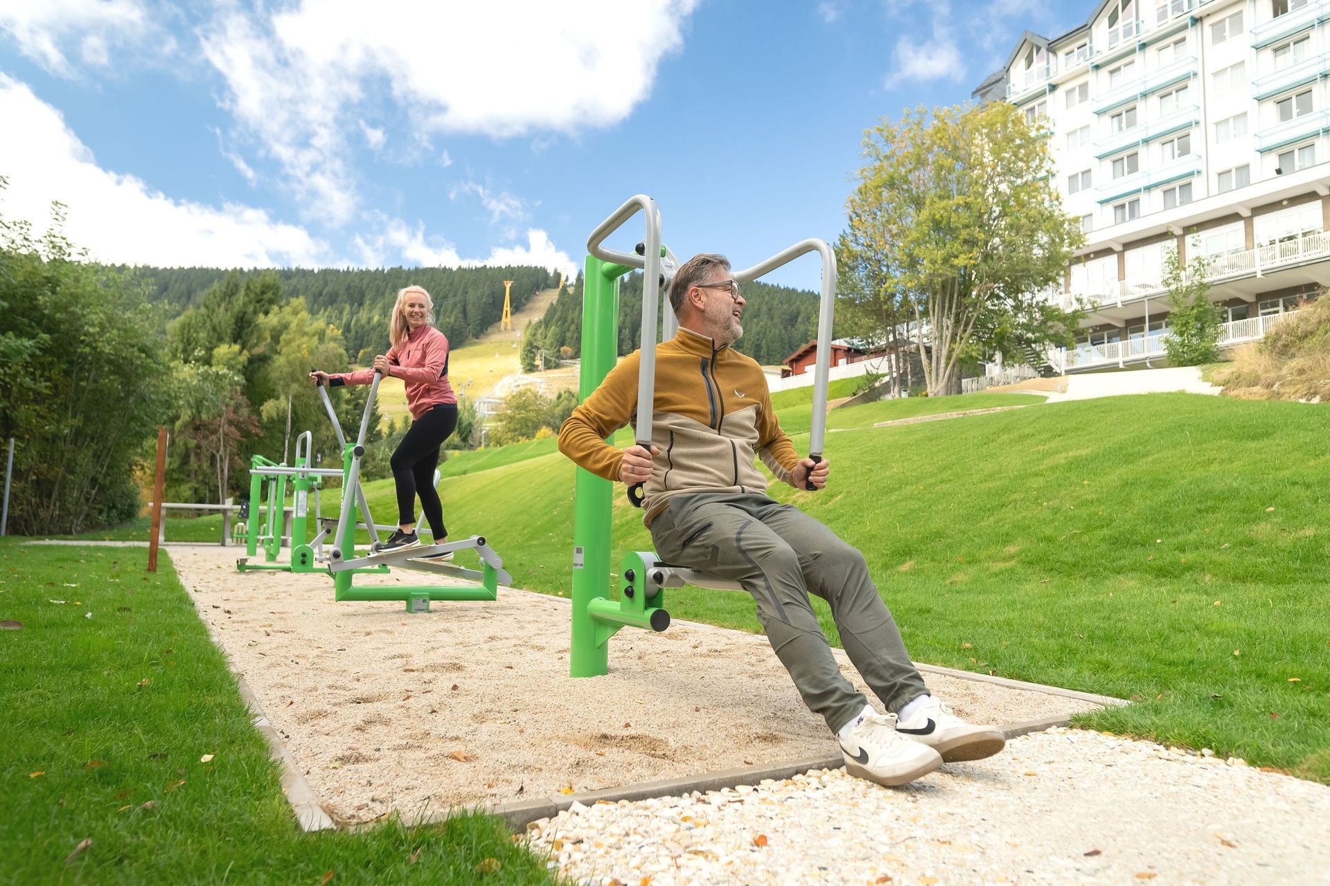 Zwei Personen benutzen Fitnessgeräte im Freien in einem Park mit Rasenfläche; im Hintergrund sind Wohnhäuser zu sehen.