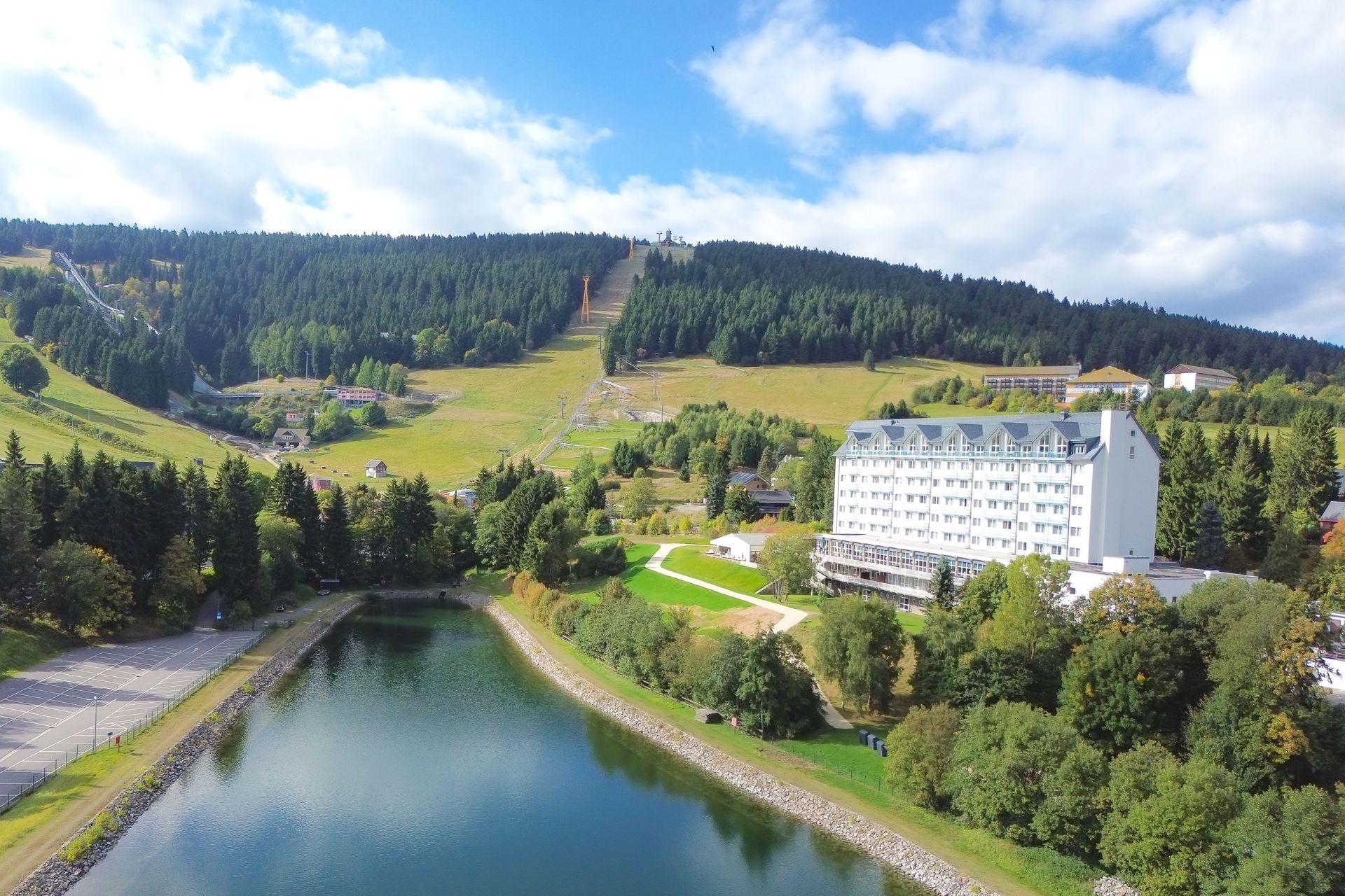 Großes weißes Hotel neben einem See und einem grünen Hügel mit einer Skisprungschanze, unter blauem Himmel.