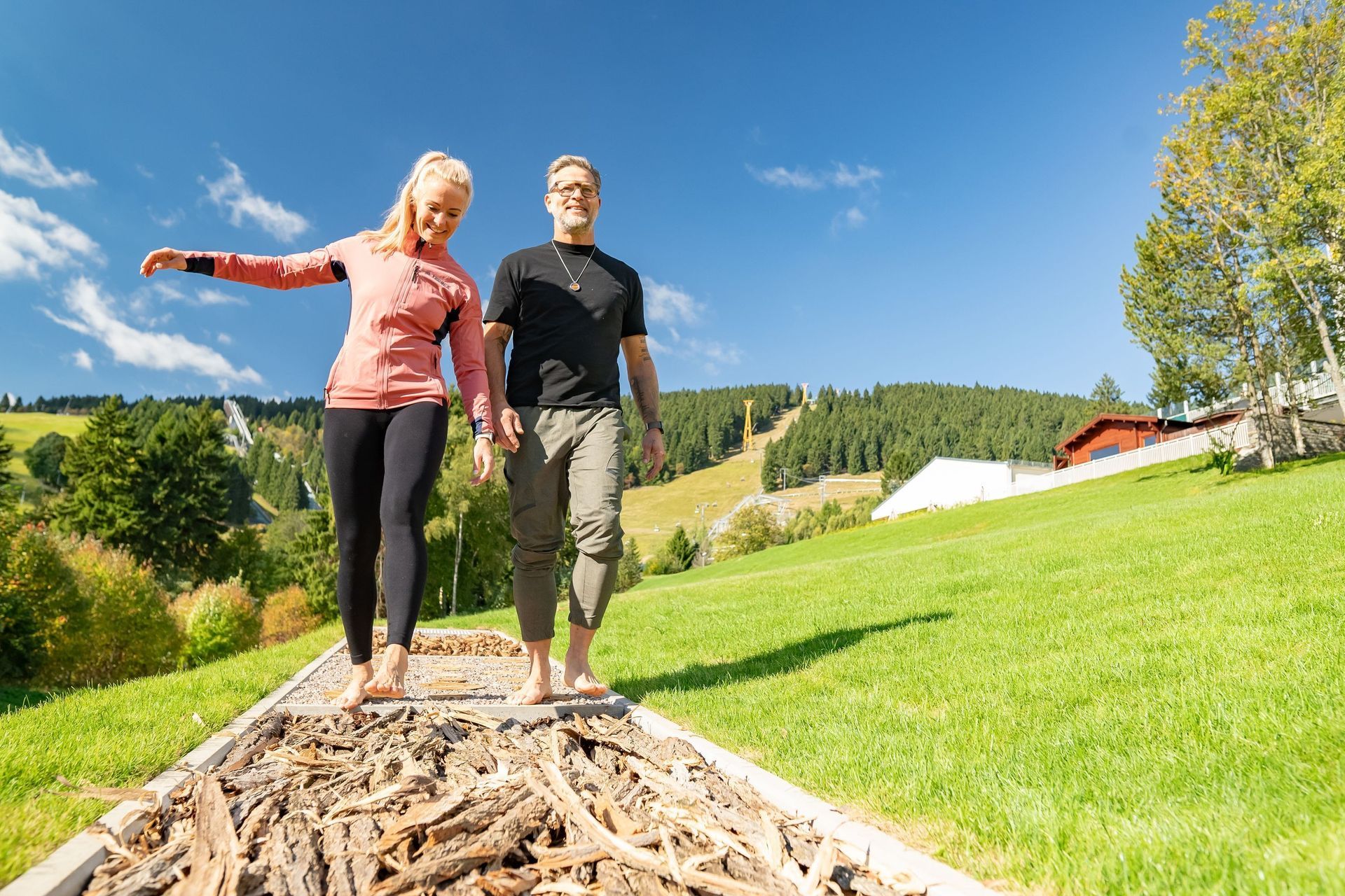 Zwei Personen spazieren auf einem Holzsteg in einer sonnigen, grasbewachsenen Hügellandschaft